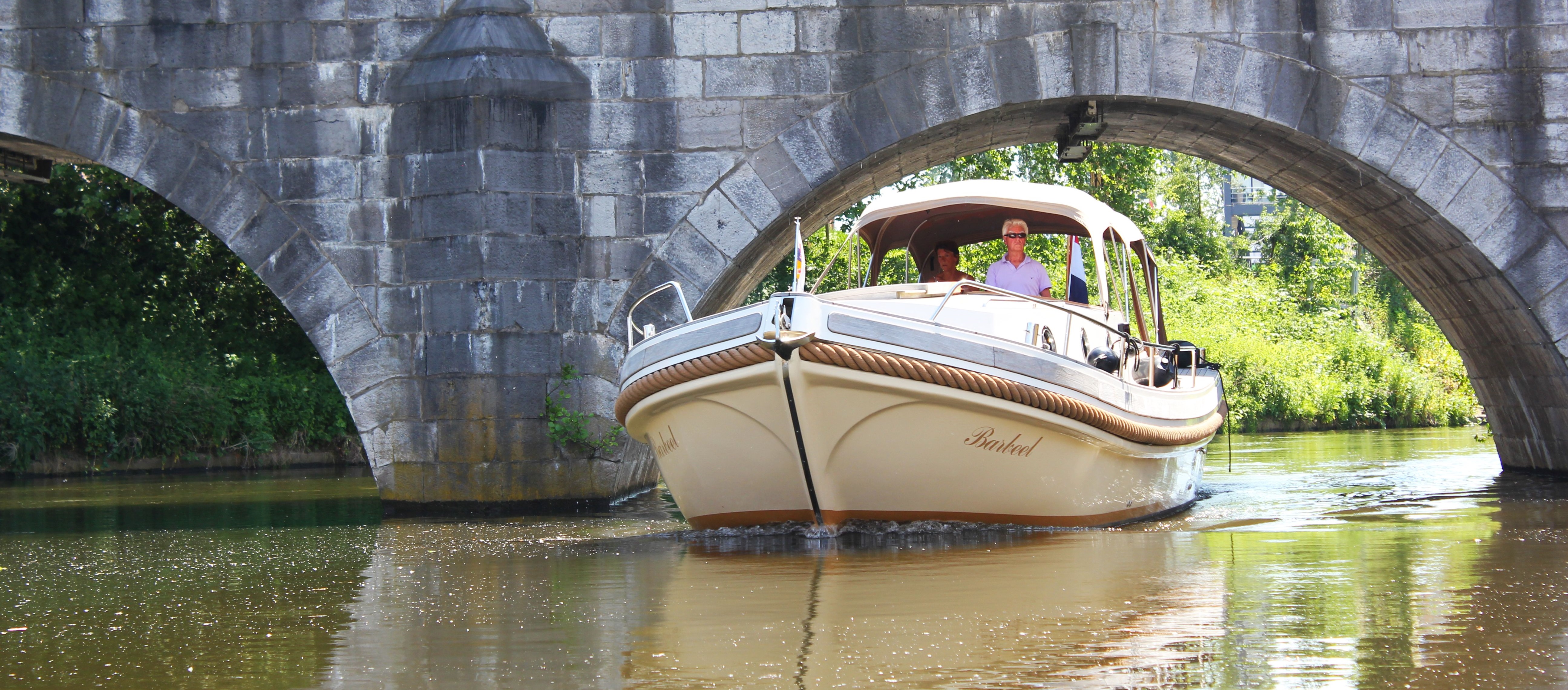 Sloep vaart onder een oude stenen brug op de Maasplassen, met mensen aan boord die genieten van een ontspannen vaartocht.