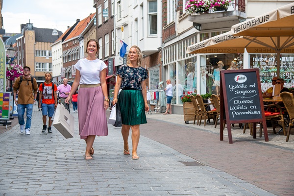 Two women walk with shopping bags through downtown Roermond, past shops and a terrace with a menu board.