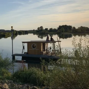 Twee mensen op een houten vlot met overnachtingshut, dobberend op de Maasplassen bij zonsondergang, omringd door groen en rust.