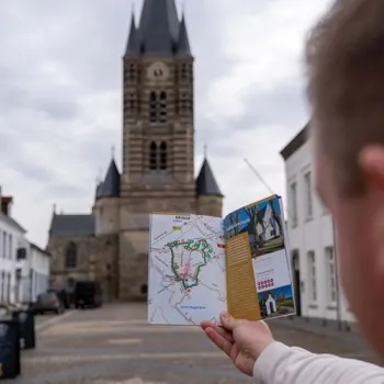 Bezoeker houdt een wandelkaart vast op een plein in Thorn, met de Abdijkerk en witte huizen op de achtergrond.