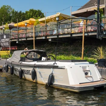 Sloep van Sloephuren-Limburg.nl ligt aangemeerd aan het water, met een terras en gele parasols op de achtergrond.